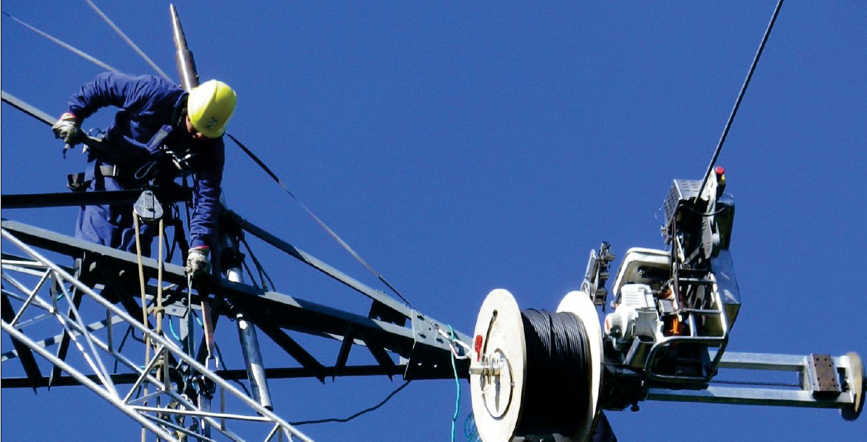 Operario en torre de transmisión con equipo de tendido y bobina de cable